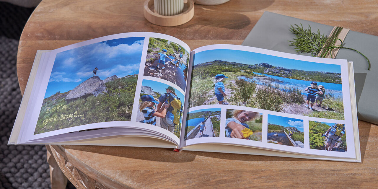 Auf einem Tisch aus Holz liegen ein aufgeschlagenes CEWE FOTOBUCH, zwei Kerzen und ein Geschenk. Auf der Doppelseite sind Fotos von einer Familie beim Wandern in den Bergen zu sehen.