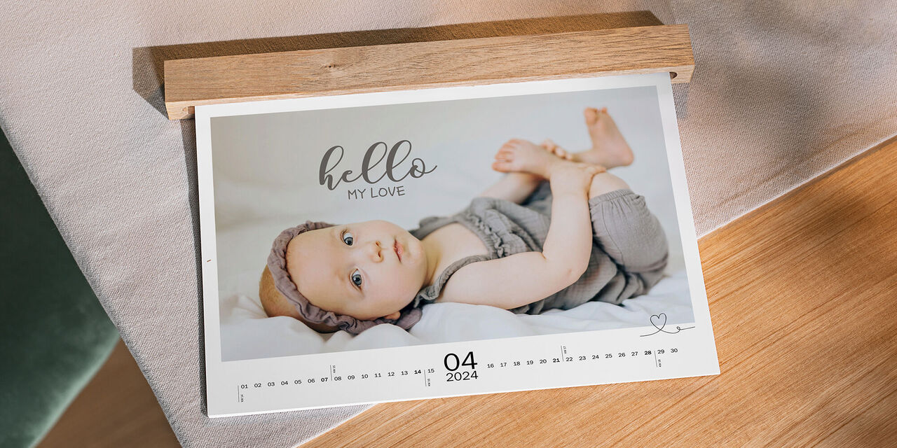 Auf einem Tisch aus Holz liegt ein Wandkalender mit Babymotiven im Querformat. Er zeigt den Monat April und hat eine Holzleiste, die die Ringbindung verdeckt.