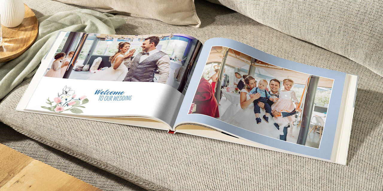 Auf einem hellen Sofa liegt ein aufgeschlagenes CEWE FOTOBUCH. Auf der linken Seite steht "Welcome to our Wedding" und man sieht das Brautpaar tanzen. Auf der rechten Seite sieht man das Brautpaar mit ihren Kindern.