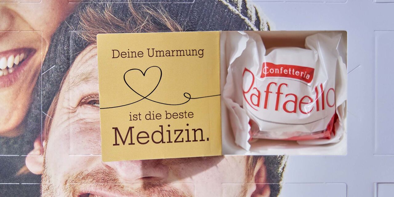 Auf der Innenseite eines geöffneten Adventskalendertürchens steht der Spruch “Deine Umarmung ist die beste Medizin”, er ist verziert mit einem Herz. Daneben steckt ein Raffaello.